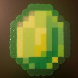 May include: A green, yellow, and light green pixelated design made of small plastic beads. The design is in the shape of a square with rounded corners.