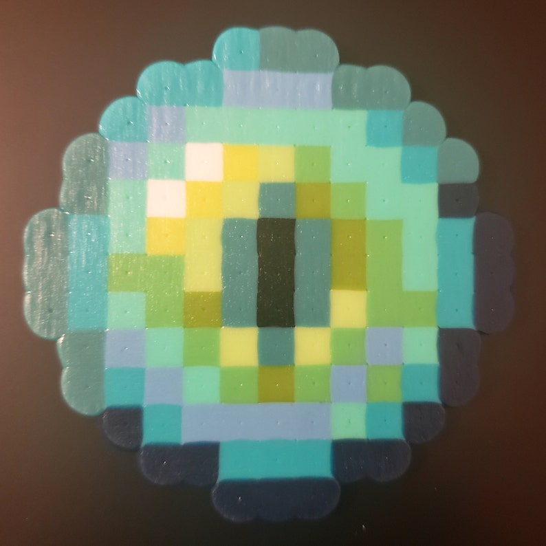 May include: A green, blue, and yellow pixelated eye with a black pupil. The eye is made of small, round beads.