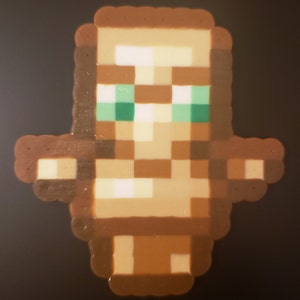May include: A brown and beige pixelated figure with green eyes. The figure is made of small beads and is a representation of a character from the video game Minecraft.