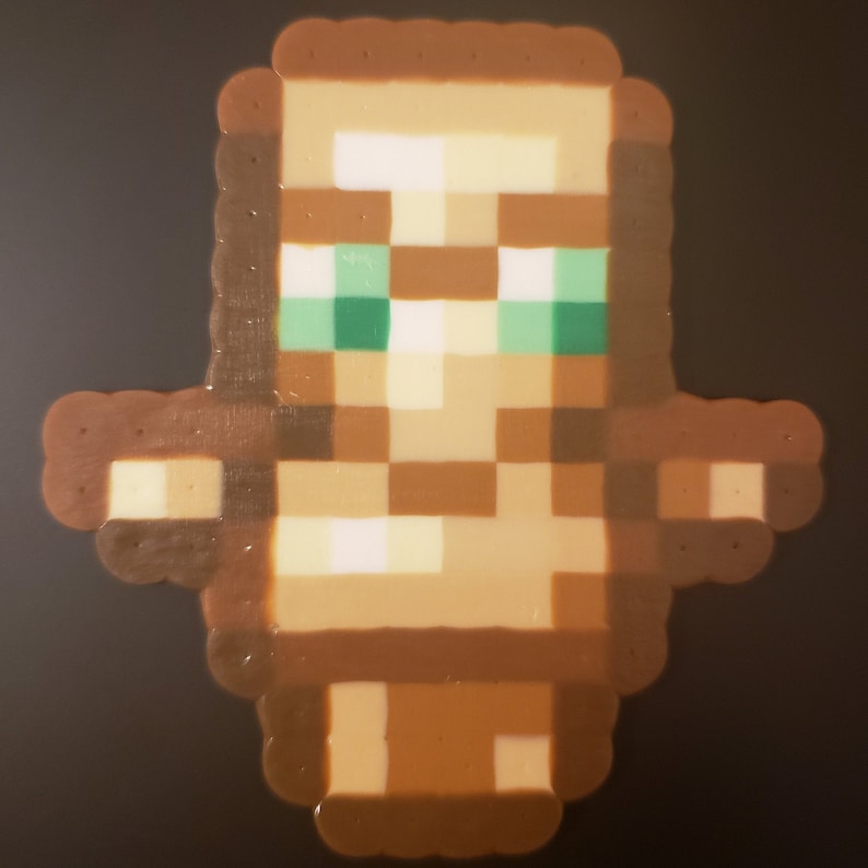 May include: A brown and beige pixelated figure with green eyes. The figure is made of small beads and is a representation of a character from the video game Minecraft.