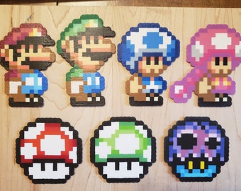 May include: A collection of pixel art characters from the Super Mario video game series. The characters include Mario, Luigi, Toad, and Toadette, along with three different colored mushrooms. The figures are made of colorful plastic beads.