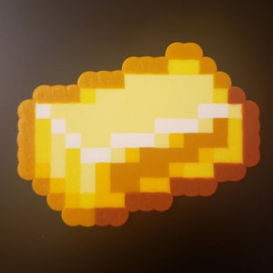 May include: A gold-colored pixelated gold bar made of plastic beads. The bar is shaped like a traditional gold bar with rounded edges. The beads are arranged in a pixelated pattern, creating a retro video game aesthetic.