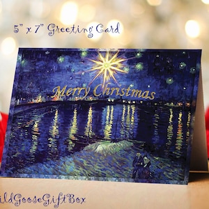 May include: A 5" x 7" Christmas greeting card featuring a starry night scene with the words "Merry Christmas" in gold. The card has a painting-like style with a dark blue and gold color scheme. The WildGooseGiftBox logo is at the bottom.