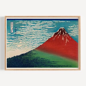 May include: A woodblock print of Mount Fuji, a famous Japanese mountain, with a red peak and a blue sky with white clouds. The print is framed in a light wood frame.