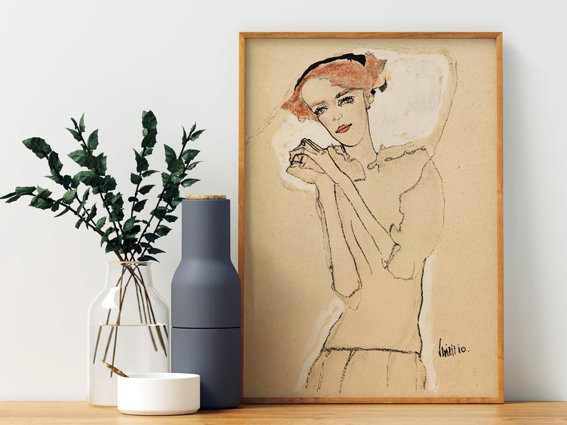 May include: A framed black and white line drawing of a woman with red hair wearing a white collar. The woman is looking down with her hands clasped in front of her. The drawing is on a beige background and signed "Klimt 10."