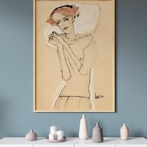 May include: A framed black and white line drawing of a woman with red hair wearing a white dress. The drawing is on a beige background and the artist's signature is visible in the bottom right corner.