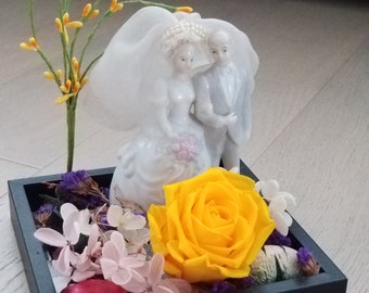 May include: A black square frame with a white porcelain figurine of a bride and groom, a yellow rose, and other dried flowers.