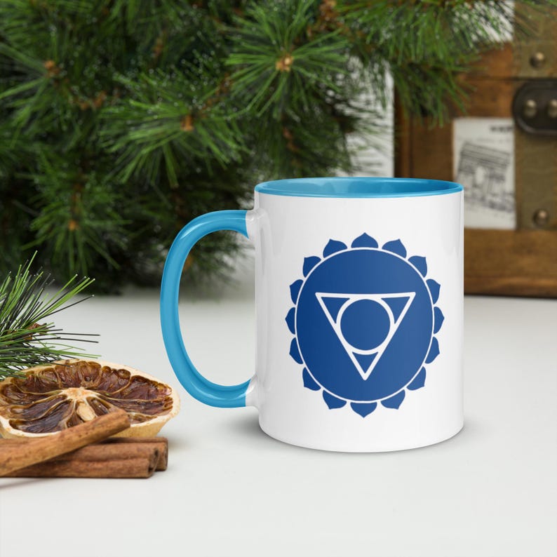 以下が含まれることがあります： 青い縁と取っ手を持つ白いセラミックマグカップ。マグカップには、コミュニケーションと表現に関連付けられている、第5チャクラである喉のチャクラの青と白のグラフィックが描かれています。