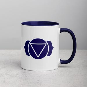 Op de afbeelding: Een witte keramische mok met een donkerblauwe rand en handvat. De mok heeft een blauw en wit symbool van de derde oog chakra, ook wel bekend als Ajna chakra.