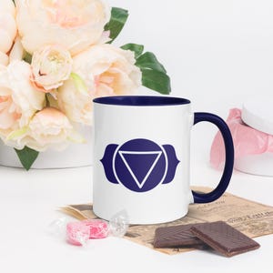 Op de afbeelding: Witte keramische mok met een blauwe rand en handvat. De mok heeft een blauw en wit symbool van de derde oog chakra, ook wel bekend als Ajna chakra.