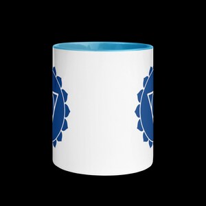 以下が含まれることがあります： 青い縁取りと両側に青いチャクラのシンボルが描かれた白いセラミック製のマグカップ。シンボルは、三角形が内側に描かれたスタイリッシュな蓮の花です。