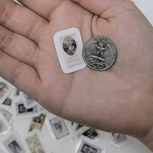 May include: A small, rectangular sticker with a religious image and text, held next to a quarter. The sticker features a blue and white illustration of figures. Other similar stickers are scattered in the background.