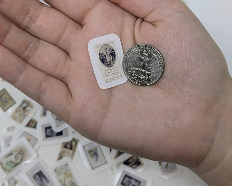 May include: A small, rectangular sticker with a religious image and text, held next to a quarter. The sticker features a blue and white illustration of figures. Other similar stickers are scattered in the background.