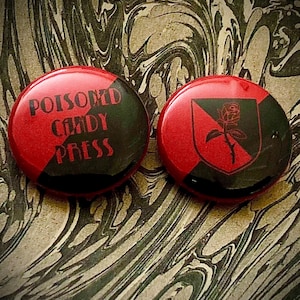 Op de afbeelding: Twee rode en zwarte ronde buttons. Eén button heeft de tekst "POISONED CANDY PRESS" in het groen. De andere button heeft een zwart schild met een roosafbeelding. De buttons liggen op een gemarmerde achtergrond.