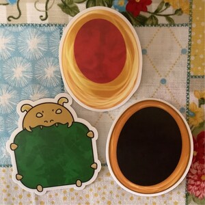 May include: Three round stickers with different designs. The first sticker is gold with a red center. The second sticker is green with a cartoon creature with a yellow head. The third sticker is gold with a black center.