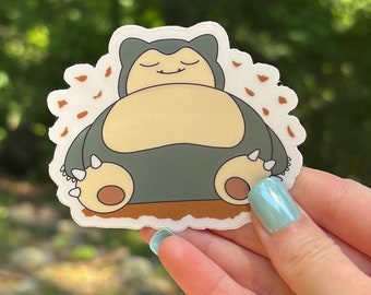 May include: A sticker featuring a cartoon Snorlax character in a sleeping pose. The sticker has a white border and is primarily colored in shades of blue-gray, tan, and brown. The background is blurred green.