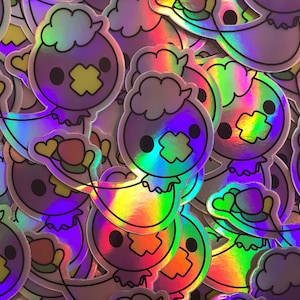 May include: A pile of iridescent holographic stickers featuring a cartoon character with a fluffy white head, a purple body, and a yellow X over its mouth. The stickers are in various positions and overlapping each other.