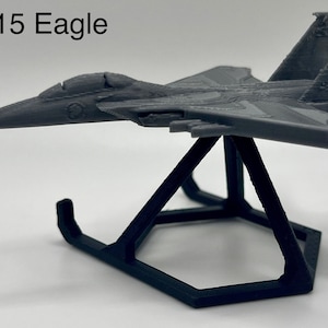 Puede incluir: Maqueta gris de un avión F-15 Eagle sobre un soporte negro. El modelo tiene alas, aletas de cola y una cabina detalladas. El texto "F-15 Eagle" es visible encima del modelo. El soporte tiene forma geométrica.