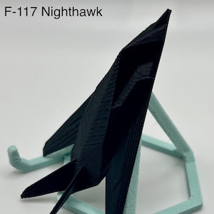 Puede incluir: Un modelo de avión de combate furtivo F-117 Nighthawk impreso en 3D en negro sobre un soporte hexagonal azul claro.
