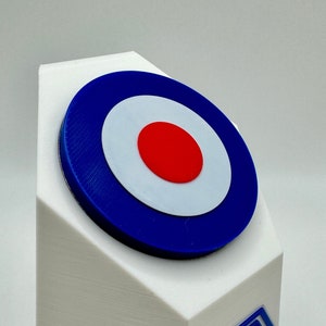 May include: A white 3D printed object with a blue and red target design on top and a blue 3D logo on the side.