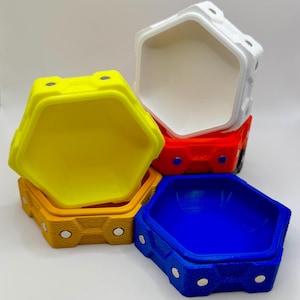 May include: A collection of brightly coloured, hexagonal containers in yellow, orange, red, white, and blue. Each container has a textured exterior and a smooth interior. The containers are designed with a geometric shape and are stacked together.