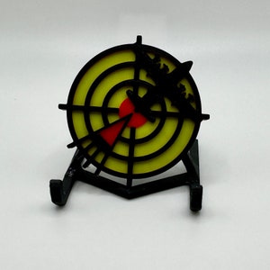 May include: A yellow and black target with a red bullseye, mounted on a black stand. The target has a black silhouette of a plane flying into it.