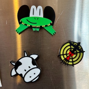 May include: Three refrigerator magnets: a green frog with black ears, a black and white cow, and a yellow target with a black airplane.