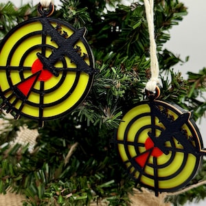 May include: Two Christmas tree decorations with a target design. Each decoration features a yellow bullseye with a red centre and a black airplane silhouette. The decorations are hanging on a Christmas tree.