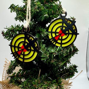 May include: Two Christmas tree ornaments with a target design. Each ornament features a yellow bullseye with black concentric circles and a red center. A black arrow shape is on top. The ornaments hang from a white string.