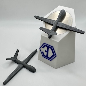 Puede incluir: Dos modelos de drones impresos en 3D de color gris. El dron más grande está en un soporte blanco con un logotipo 3D azul. El dron más pequeño está acostado sobre una superficie gris.