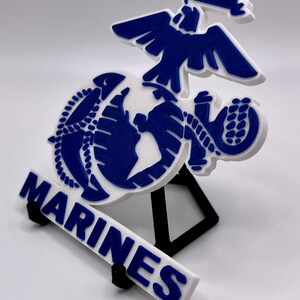 May include: A white 3D printed model of the United States Marine Corps emblem. The emblem features a globe with an eagle perched on top and the word "MARINES" below. The model is mounted on a black stand.