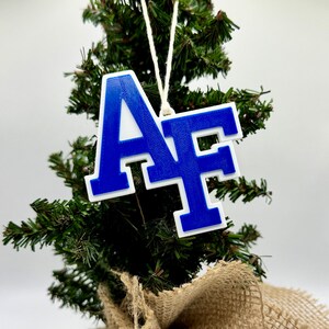 May include: A small artificial Christmas tree with a burlap base. A blue and white ornament with the letters "AF" hangs from the tree. The tree is decorated with a white string.