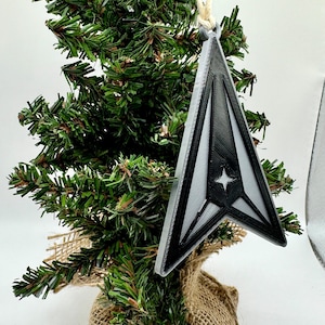 Pode incluir: Um ornamento triangular preto e prateado com um desenho de estrela, pendurado numa pequena árvore de Natal. A árvore é embrulhada em serapilheira. O ornamento é suspenso por um fio natural.