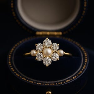 Puede incluir: Un anillo de oro con diseño floral, engastado con diamantes y perlas. El anillo presenta una perla central rodeada de seis diamantes, con perlas más pequeñas a cada lado. El anillo se presenta en un joyero de terciopelo azul.