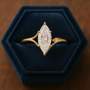 Könnte beinhalten: Ein Verlobungsring mit Diamanten im Marquiseschliff und einem goldenen Band. Der Ring hat einen großen, klaren Diamanten in einer Krappenfassung. Das Band hat zarte Details und einen kleinen Diamantakzent auf jeder Seite. Der Ring wird in einer blauen Samtringbox präsentiert.