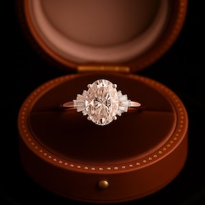 Puede incluir: Un anillo de compromiso de diamantes de talla ovalada con piedras laterales de baguette, engastado en una banda de oro rosa, descansa dentro de un joyero de cuero marr&oacute;n. El anillo es el punto focal, con la caja como tel&oacute;n de fondo.