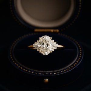 Puede incluir: Anillo de compromiso de oro con un diamante central de talla ovalada y diamantes baguette m&aacute;s peque&ntilde;os a cada lado. El anillo se presenta en una caja de terciopelo azul oscuro.