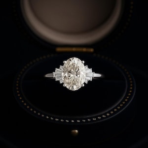 Puede incluir: Un anillo de compromiso de diamantes de talla ovalada con piedras laterales de baguette, engastado en una banda plateada. El anillo se muestra en un joyero negro con interior dorado. La piedra central grande es el punto focal.