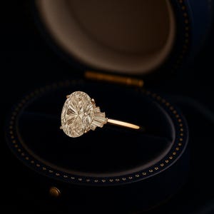 Puede incluir: Un anillo de oro con un gran diamante de talla ovalada como pieza central, engastado en una banda de oro. El diamante est&aacute; rodeado de diamantes m&aacute;s peque&ntilde;os, de talla baguette, a cada lado. El anillo se muestra en un joyero azul oscuro.
