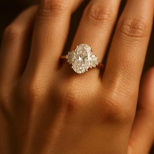 Puede incluir: Un anillo de oro con un gran diamante de talla ovalada como pieza central, flanqueado por diamantes m&aacute;s peque&ntilde;os de talla baguette. El anillo est&aacute; engarzado en una delicada banda de oro, mostrando un dise&ntilde;o cl&aacute;sico y elegante. El anillo se muestra en una mano.