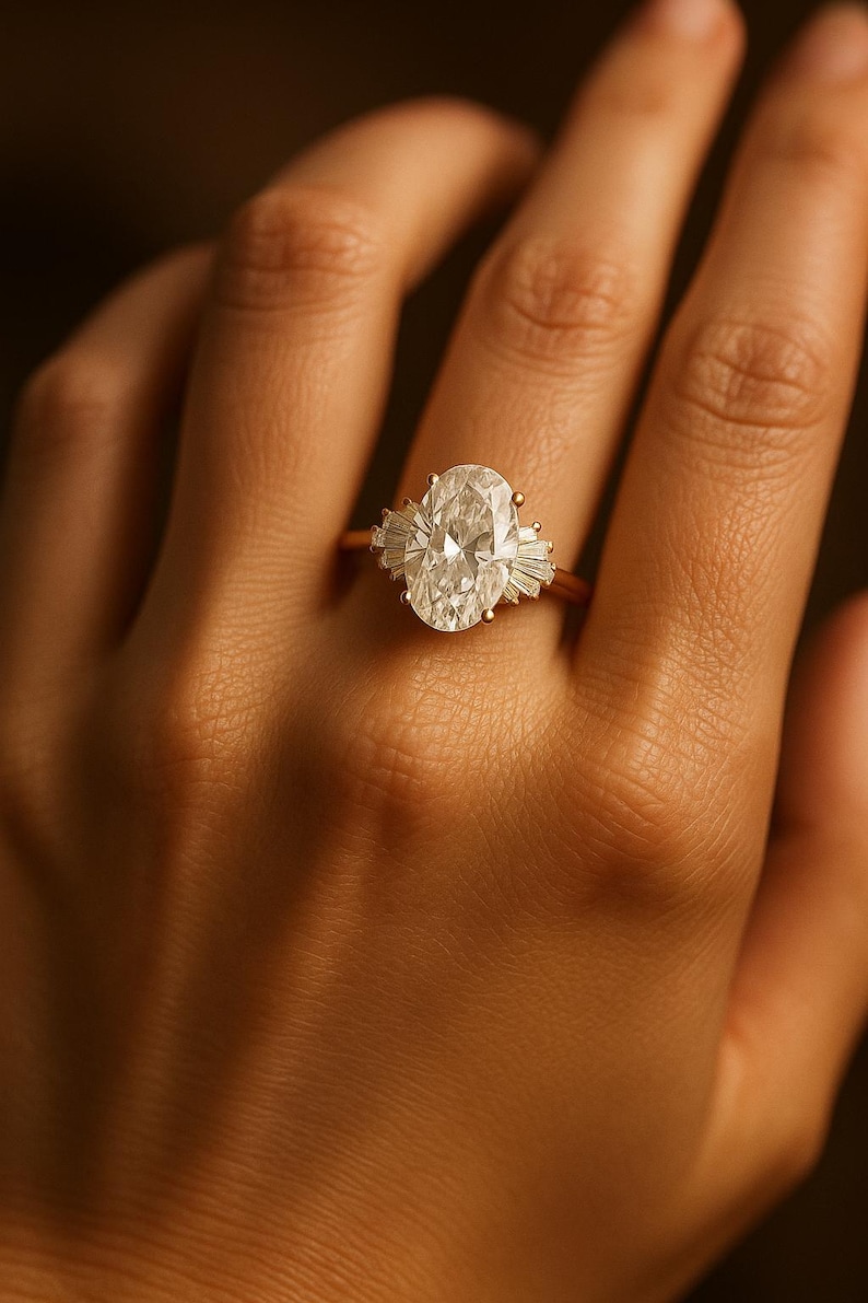 Puede incluir: Un anillo de oro con un gran diamante de talla ovalada como pieza central, flanqueado por diamantes m&aacute;s peque&ntilde;os de talla baguette. El anillo est&aacute; engarzado en una delicada banda de oro, mostrando un dise&ntilde;o cl&aacute;sico y elegante. El anillo se muestra en una mano.