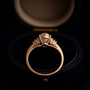 Puede incluir: Un anillo de compromiso de oro con una piedra central grande y transparente de talla redonda y piedras laterales m&aacute;s peque&ntilde;as de talla baguette. El anillo est&aacute; engastado en una banda de oro.