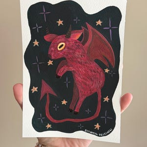 May include: A rectangular art print featuring a dark red, winged creature with horns and a pointed tail, set against a black background filled with small orange stars and purple cross-shaped sparkles. The artist name Elizabeth Deczynski is printed in white at the bottom.