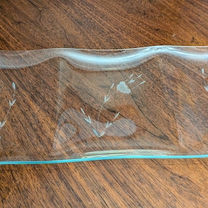 May include: A clear, rectangular glass serving dish with a slightly wavy design. The dish features etched floral and leaf patterns. The edges have a subtle blue tint. The dish is displayed on a dark wood surface.