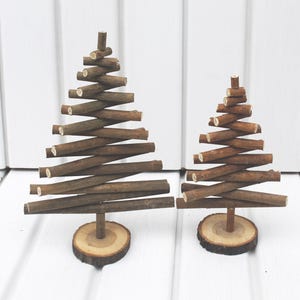 Könnte beinhalten: Zwei dekorative Weihnachtsbäume aus Holz. Die Bäume bestehen aus kleinen, braunen Ästen, die in einer dreieckigen Form angeordnet sind. Jeder Baum ist an einer kleinen, runden Holzbasis befestigt. Der Hintergrund ist weiß.