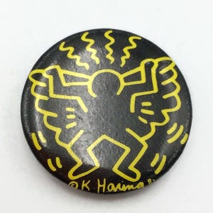 May include: A round, black button featuring a yellow graphic of a stylized figure with outstretched arms and wings, surrounded by radiating lines. The button has the text "© K Haring 87" in yellow.