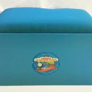 May include: A teal storage bench with a padded top. The bench has a wooden frame and a circular sticker on the front with the text "Thomas The Tank Engine & Friends". The bench has small teal fabric loops on the sides.
