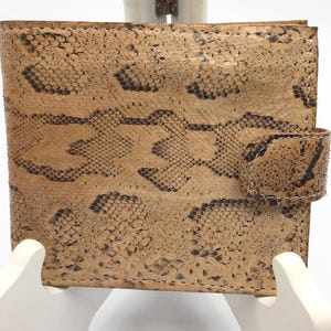 May include: A tan and brown leather wallet with a snakeskin pattern. The wallet has a rectangular shape with a snap closure. The exterior features a detailed snakeskin texture, with dark brown and tan scales. The wallet is displayed on a white stand.