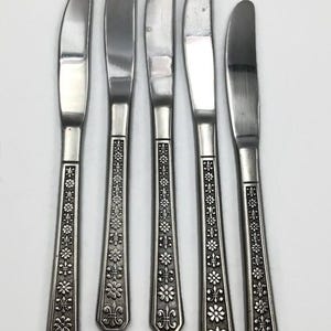 May include: Five vintage butter knives with a silver finish. The handles feature an ornate floral and fleur-de-lis design. The blades are curved and slightly rounded.