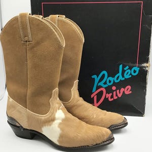Könnte beinhalten: Ein Paar hellbraune Cowboy-Stiefel mit einem hellbraunen Wildleder-Obermaterial und einem Kuhfellfuß mit weißen Flecken. Die Stiefel haben eine spitze Zehenpartie und einen schwarzen Absatz. Auf der Schachtel hinter den Stiefeln steht "Rodeo Drive".